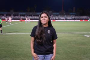 A teenage girl stands smiling on a soccer field at night, wearing a Las Vegas Lights FC t-shirt and jeans, with players and spectators visible in the background under stadium lighting.