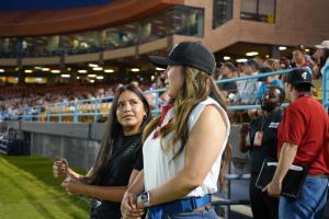 A teenage girl wearing a Las Vegas Lights FC shirt stands on the sidelines of a stadium, looking up at a woman in a black cap and white top with a red scarf. Fans fill the stands in the background during an evening soccer match.