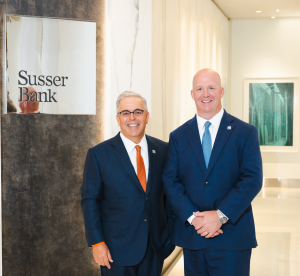 Sam L. Susser, Chairman and CEO of Susser Bank, and Carl Cravens, President and Chief Banking Officer of Susser Bank, stand side by side in professional attire in the bank’s modern lobby. A polished wall sign featuring the Susser Bank logo is visible to t