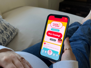 A pregnant woman in jeans holds a phone displaying the "Count the Kicks" app, with options to start counting and surveys, conveying calm anticipation.
