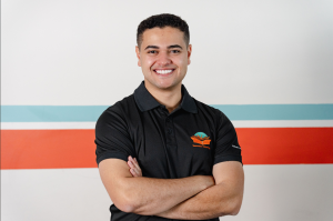Michael Black, smiling with arms crossed, wearing a Success Tutoring shirt, stands in front of a wall featuring teal, white and red stripes.