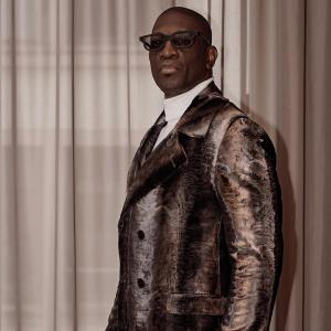 Michael Ameer wearing a velvet-patterned brown suit and black glasses, standing confidently in front of soft beige curtains in a formal press photo