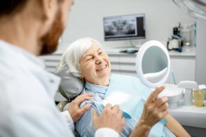 A senior woman smiles in the mirror while admiring her new dentures as the dentist observes.