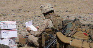U.S. Marine in full gear reading a handwritten letter next to several USPS mail boxes in a desert deployment zone.