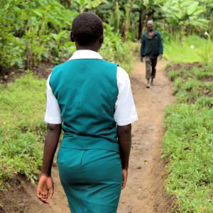girl in school uniform walks away from camera along a rural path through a jungle, coming towards her is a man, whose image is out of focus