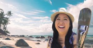Photo of Hawaii artist Jan Tetsutani looks happy wearing a hat and holding her paintbrush at a sunny beach in Hawaii